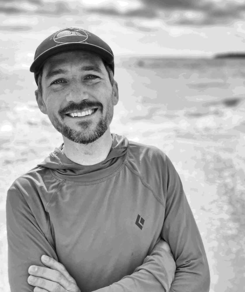 Black and white portrait of Ryland in a cap, smiling on a beach